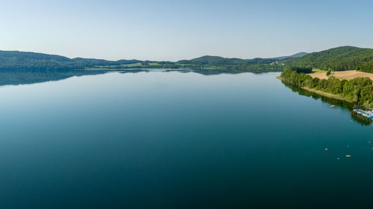 A tranquil lake with clear water and green shores. In the background, there are gentle hills and some boats visible.