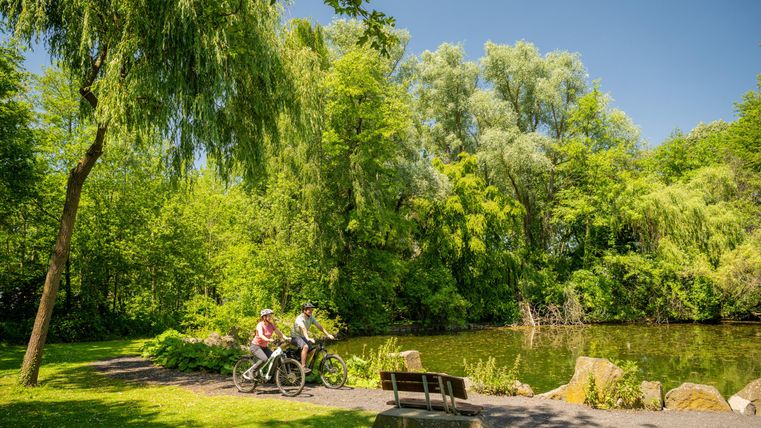 Zwei Personen fahren mit Fahrrädern entlang eines ruhigen Gewässers in einer grünen Parklandschaft. Die Umgebung ist üppig bewachsen mit Bäumen und Sträuchern.