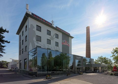 Ein modernes Hotelgebäude mit großen Glasfenstern und einem historischen Schornstein. Die Fassade zeigt eine stilisierte Figur und die Sonne scheint hell am Himmel.