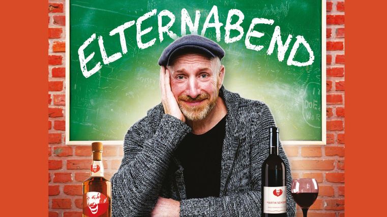 Ein Mann sitzt lächelnd vor einer Tafel mit der Aufschrift "Elternabend". Neben ihm stehen Flaschen Wein und ein Glas.