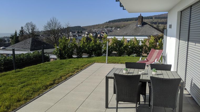 Eine sonnige Terrasse mit einem Tisch und Stühlen. Im Hintergrund ist eine grüne Wiese und sanfte Hügel zu sehen.