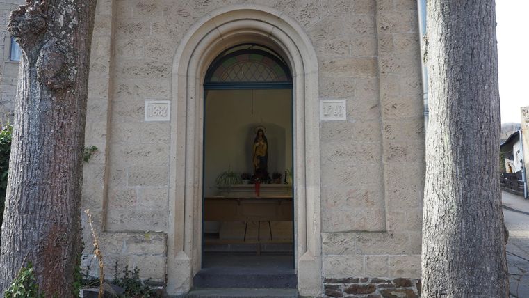 Ein Eingang zu einer kleinen Kapelle mit einem dekorativen Bogen. Im Inneren ist eine Statue zu sehen, umgeben von einem schlichten, ruhigen Raum.