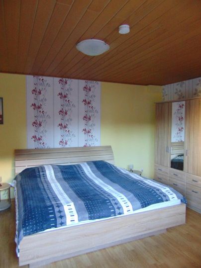 A cozy bedroom with a wooden bed and colorful bedding. The walls are light yellow, and there is a wooden wardrobe.