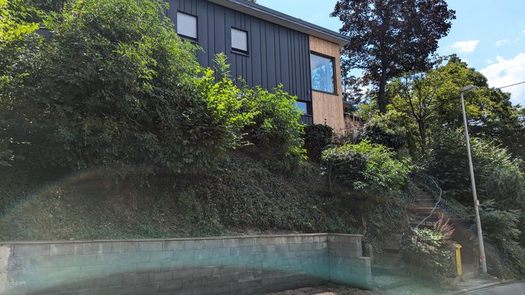 Ein modernes Haus liegt auf einem Hügel, umgeben von viel grünem Gestrüpp. Die Fassade des Hauses ist schwarz mit Holzakzenten.