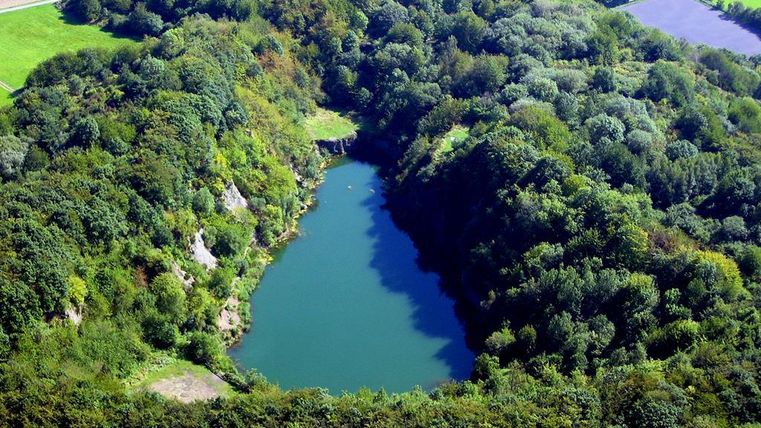 Ein von Bäumen umgebenes, blaues Gewässer in einer grünen Landschaft. Der See ist von hügeligem Gelände und Wäldern umgeben.