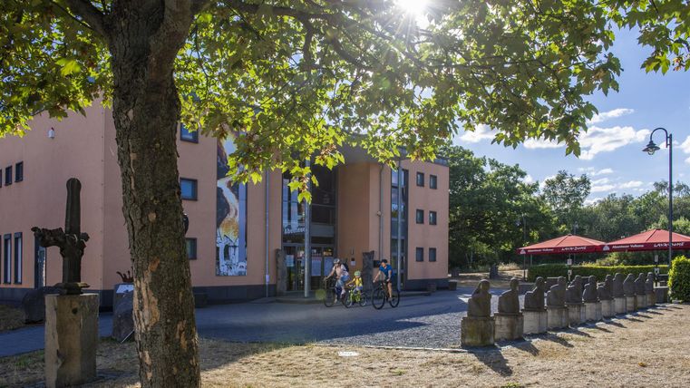 Ein modernes Gebäude umgeben von Bäumen und Sonnenschein. Im Vordergrund sind Menschen zu sehen, die entspannt eine Terrasse nutzen.