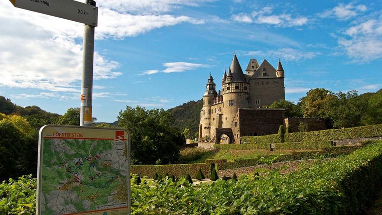 Eine malerische Burg umgeben von grünen Landschaften und Himmel mit einigen Wolken. Im Vordergrund sind ein Wegweiser und eine Karte sichtbar.