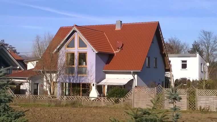 Ein modernes, zwei Geschosse umfassendes Haus mit rotem Dach und großen Fenstern. Der Garten ist von einem Holzzaun umgeben und es gibt einige Bäume in der Nähe.
