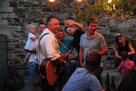 Eine fröhliche Gruppe von Menschen feiert im Freien, während ein Musiker mit einer Gitarre spielt. Lichterketten sorgen für eine gemütliche Atmosphäre.