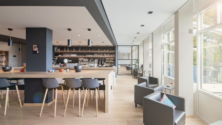 Ein modernes Café mit einem hellen, einladenden Innenraum. Im Hintergrund sieht man eine lange Theke und gemütliche Sitzmöglichkeiten.