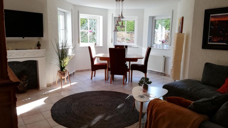 A bright living room with a round rug, a sofa, and a dining table. Large windows provide plenty of daylight and a welcoming atmosphere.