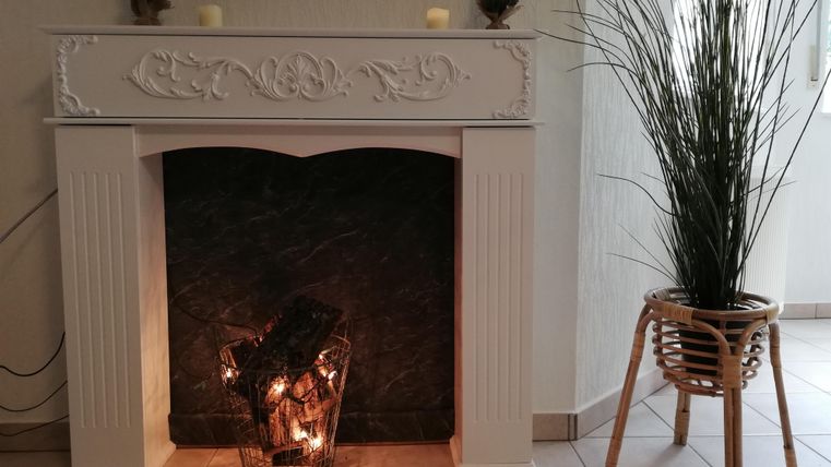 A cozy fireplace with decorative candles and a modern mantel. Next to the fireplace stands a planter with tall, green plants.