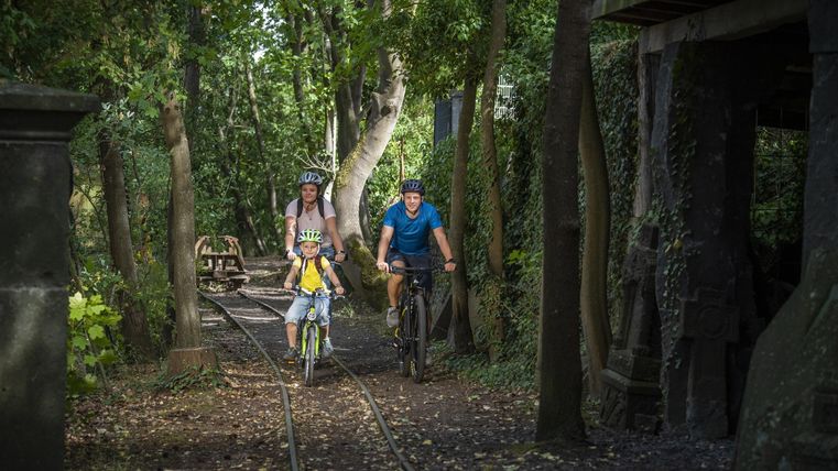 A family is riding bicycles on a narrow path through a wooded area. The surroundings are green and inviting, ideal for outings in nature.
