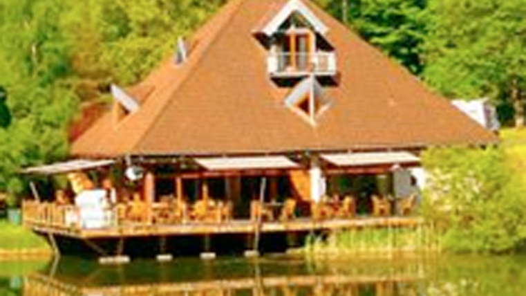 Ein gemütliches Holzhaus am Ufer eines Sees, umgeben von Bäumen. Die Terrasse ist mit Stühlen und Tischen ausgestattet, die einen Blick auf das Wasser bieten.