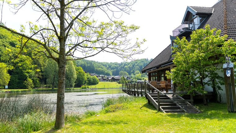A picturesque landscape with a lake and beautiful accommodation. Surrounded by green trees and gentle hills, the place offers a tranquil atmosphere.
