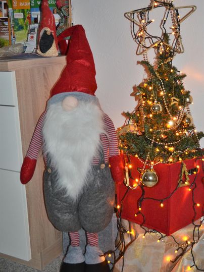 Ein fröhlicher Weihnachtswichtel mit einem roten Hut steht neben einem geschmückten Weihnachtsbaum. Der Baum ist mit Lichtern und goldenen Kugeln verziert.