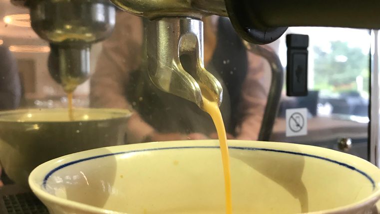 A freshly brewed coffee flows into a white cup. In the background, the details of the coffee machine are visible.