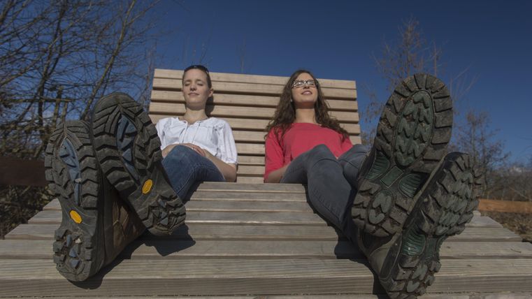 Two women are sitting relaxed on a wooden lounge chair. You can see their legs and shoes as they enjoy the sun.