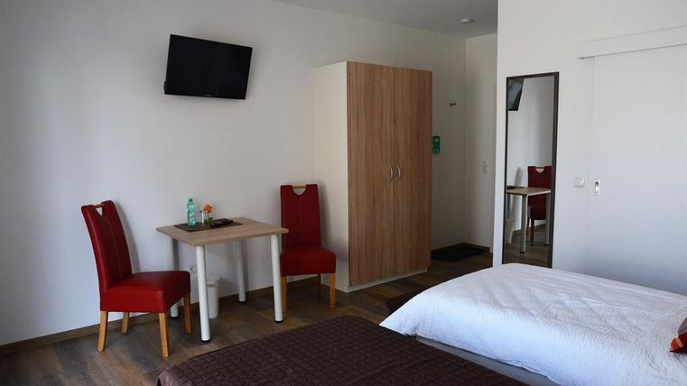 Ein einfaches Zimmer mit zwei Betten, einem Tisch und roten Stühlen. Ein Fernseher hängt an der Wand und ein Kleiderschrank steht in der Ecke.