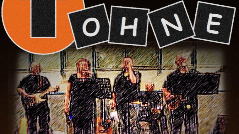 Eine Band spielt auf der Bühne, bestehend aus mehreren Musikern. Im Hintergrund ist ein großes, orangefarbenes Logo mit einem Pfeil zu sehen.