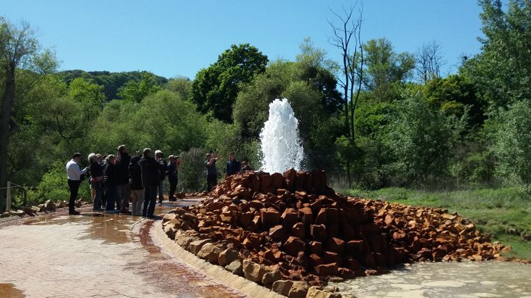 Ein sprudelnder Brunnen umgeben von Steinen und Bäumen. Eine Gruppe von Menschen steht in der Nähe und genießt die Aussicht.