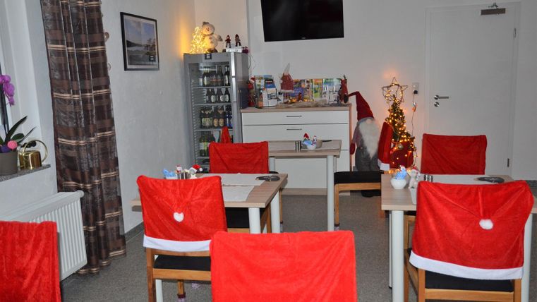 Ein gemütlicher Raum mit Tischen und Stühlen, die mit roten Weihnachtsüberwürfen dekoriert sind. Im Hintergrund steht ein kleiner Weihnachtsbaum und festliche Dekorationen sind sichtbar.