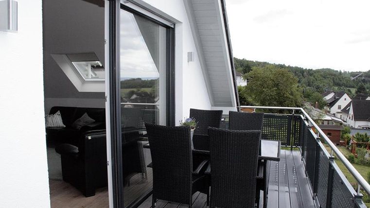 Ein moderner Balkon mit einem Essbereich aus schwarzem Rattanmöbel. Im Hintergrund ist eine grüne Landschaft und einige Wohnhäuser zu sehen.