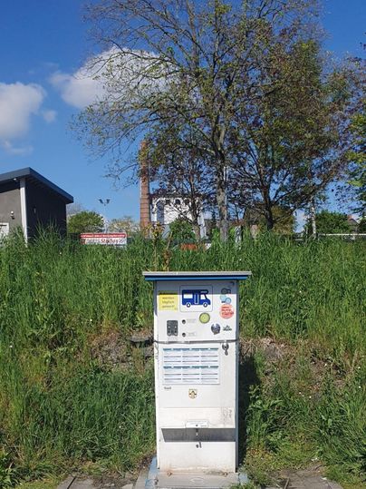 A charging station for electric cars is in front of tall grass and a blue sky. In the background, some buildings and trees are visible.
