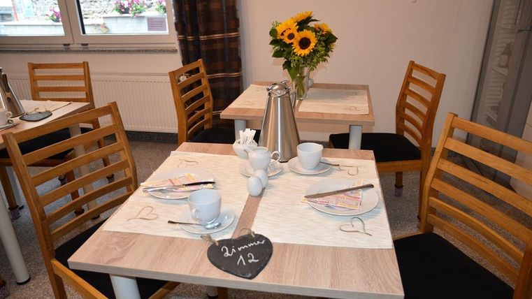 Ein gemütlicher Tisch mit Holzstühlen in einem hellen Raum. Auf dem Tisch stehen eine Kaffeekanne, Tassen und ein Strauß Sonnenblumen.