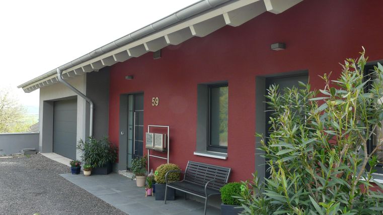 A modern house with a red facade and a green front yard. A wooden seating area is placed in front of the entrance.