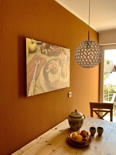 Ein gemütlicher Essbereich mit einer Holztisch und einer großen Pendelleuchte. An der Wand hängt ein Bild von leckeren Speisen, und auf dem Tisch stehen eine dekorative Schale mit Obst und ein kleiner Behälter.